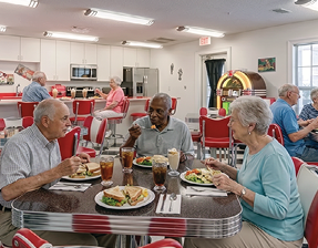 An elderly woman is sitting at a table with a plate of food in front of her. She is smiling and appears to be enjoying her meal. There are other people in the background, and a potted plant is visible in the room. AI generated content